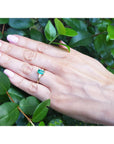 Hand wearing a ring with a natural colombian emerald against a green leafy background.