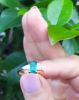 Hand holding a Colombian emerald ring with a green gemstone against a leafy background.