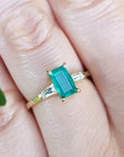 Gold ring with a natural emerald on a finger, with a blurred green leaf in the background.