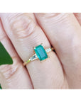 Gold ring with a natural emerald on a finger, with a blurred green leaf in the background.
