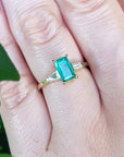 Gold ring with a real emerald on a person's finger, with a blurred green leaf in the background.