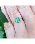 Gold ring with a real emerald on a person's finger, with a blurred green leaf in the background.
