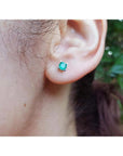 Close-up of an ear wearing a emerald stud earring with a blurred natural background.