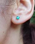 Close-up of an ear wearing a emerald stud earring with a blurred natural background.