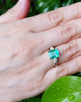 Hand wearing an emerald ring with a green leafy background.