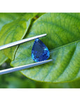 3.67 ct. deep blue pear-cut natural sapphire held by tweezers against green leaves.