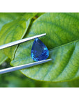 3.67 ct. deep blue pear-cut natural sapphire held by tweezers against green leaves.