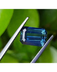 5.05 ct. GIA certified deep blue-green emerald cut sapphire held by tweezers.