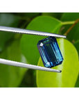 GIA certified 5.05 ct deep blue-green emerald cut sapphire held by tweezers against green leaves.
