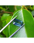 Deep blue-green emerald cut sapphire held by tweezers on a green leaf.