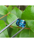 4.52 ct untreated deep blue-green sapphire, emerald cut, held by tweezers against green leaves.