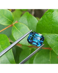4.52 ct untreated deep blue-green sapphire, emerald cut, held by tweezers against green leaves.