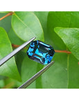 4.52 ct untreated deep blue-green emerald cut natural sapphire held by tweezers against green leaves.