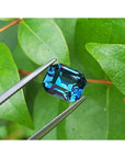 4.52 ct untreated deep blue-green emerald cut natural sapphire held by tweezers against green leaves.