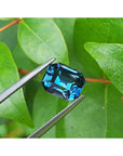 4.52 ct untreated deep blue-green emerald cut natural sapphire held by tweezers against green leaves.