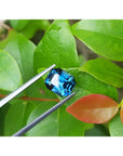 4.52 ct. untreated deep blue-green emerald cut sapphire held by tweezers against green leaves.