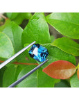 4.52 ct. untreated deep blue-green emerald cut sapphire held by tweezers against green leaves.