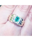 A men's 14k gold ring featuring a square natural Colombian emerald, accented with diamonds, worn between the fingers.