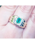 A men's 14k gold ring featuring a square natural Colombian emerald, accented with diamonds, worn between the fingers.