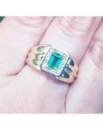A men's 14k gold ring featuring a square natural Colombian emerald, accented with diamonds, worn between the fingers.