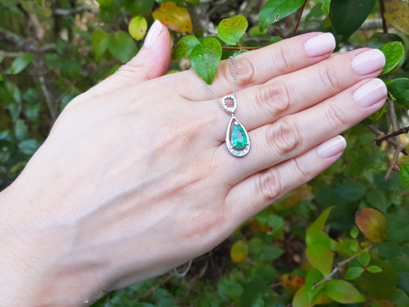 Hand displaying a white gold pendant with a natural emerald against a natural background.