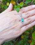 Hand displaying a white gold pendant with a natural emerald against a natural background.