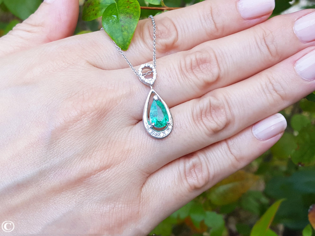 White gold necklace with a pear cut emerald held on a hand against a natural background.