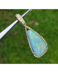 Opal pendant with gold frame held by tweezers against a blurred green background.