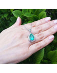 Hand holding a white gold necklace with a natural emerald pendant against a green leafy background.