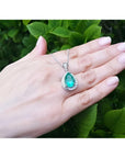 Pear-shaped natural emerald pendant with diamond halo on a 14K white gold chain, worn on a hand.