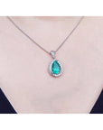 Pear-shaped natural emerald pendant with diamond halo on a 14K white gold chain, worn on a person.