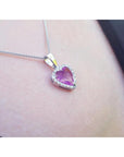 Heart-cut pink sapphire pendant with a diamond halo on an 18K white gold chain, worn on a person's neck.