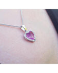 Heart-cut pink sapphire pendant with a diamond halo on an 18K white gold chain, worn on a person's neck.