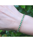 Gold bracelet with round cut natural colombian emeralds on a wrist against a green leafy background.