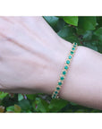 Gold bracelet with round cut natural colombian emeralds on a wrist against a green leafy background.