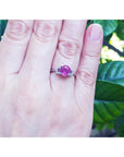 Hand wearing a ring with a pink natural ruby gemstone against a green leafy background