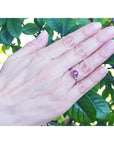 Round cut natural ruby ring with side diamonds in 14k white gold on a hand, surrounded by green leaves.