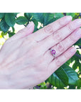 Round cut natural ruby ring with side diamonds in 14k white gold on a hand, surrounded by green leaves.