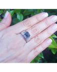 Hand wearing a white gold ring with a blue sapphire gemstone against a green leafy background