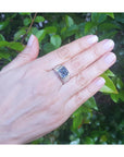 Hand wearing a white gold ring with a blue natural sapphire gemstone against a green leafy background