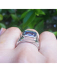 18k white gold ring with a blue natural sapphire worn on a finger against a green leafy background