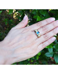 Hand wearing a gold ring with a blue natural sapphire stone against a green leafy background