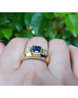 Gold ring with a blue natural sapphire and diamonds gemstone on a finger against a green leafy background