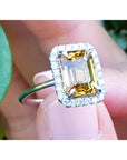 Emerald cut 4.59 ct yellow sapphire halo diamond ring in 14K white gold, worn on a finger.