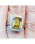 Emerald cut yellow sapphire ring with diamond halo in 14K white gold on a finger.