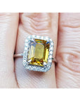 Emerald cut yellow sapphire ring with diamond halo in 14K white gold on a finger.