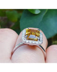 Emerald cut yellow sapphire ring with diamond halo in 14K white gold on a finger.