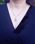 Pear cut emerald necklace with double diamond halo on 18K white gold chain, worn on a woman.