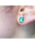 Oval natural emerald and diamond earrings in 14K white gold on an ear.