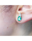 Oval natural emerald and diamond earrings in 14K white gold on an ear.